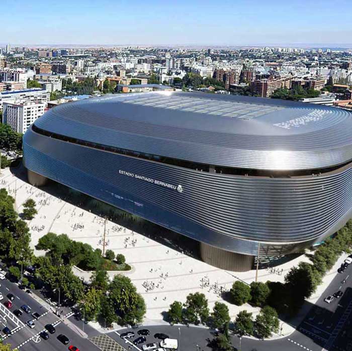 Vista aérea de un estadio moderno y elegante con un exterior metálico, rodeado de árboles y edificios urbanos. La arquitectura presenta líneas suaves y curvas y una plaza abierta y ajardinada. Se ven carreteras y coches en primer plano.