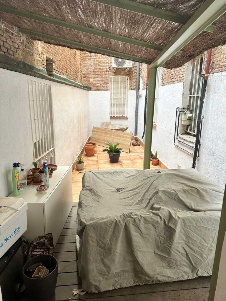 Un pequeño patio cerrado con parrilla techada, plantas en macetas y paredes de ladrillo. Los artículos de limpieza están en un armario. El suelo es de baldosas y el espacio cuenta con sombra gracias a un techo. Paredes blancas con ventanas enrejadas flanquean el espacio.
