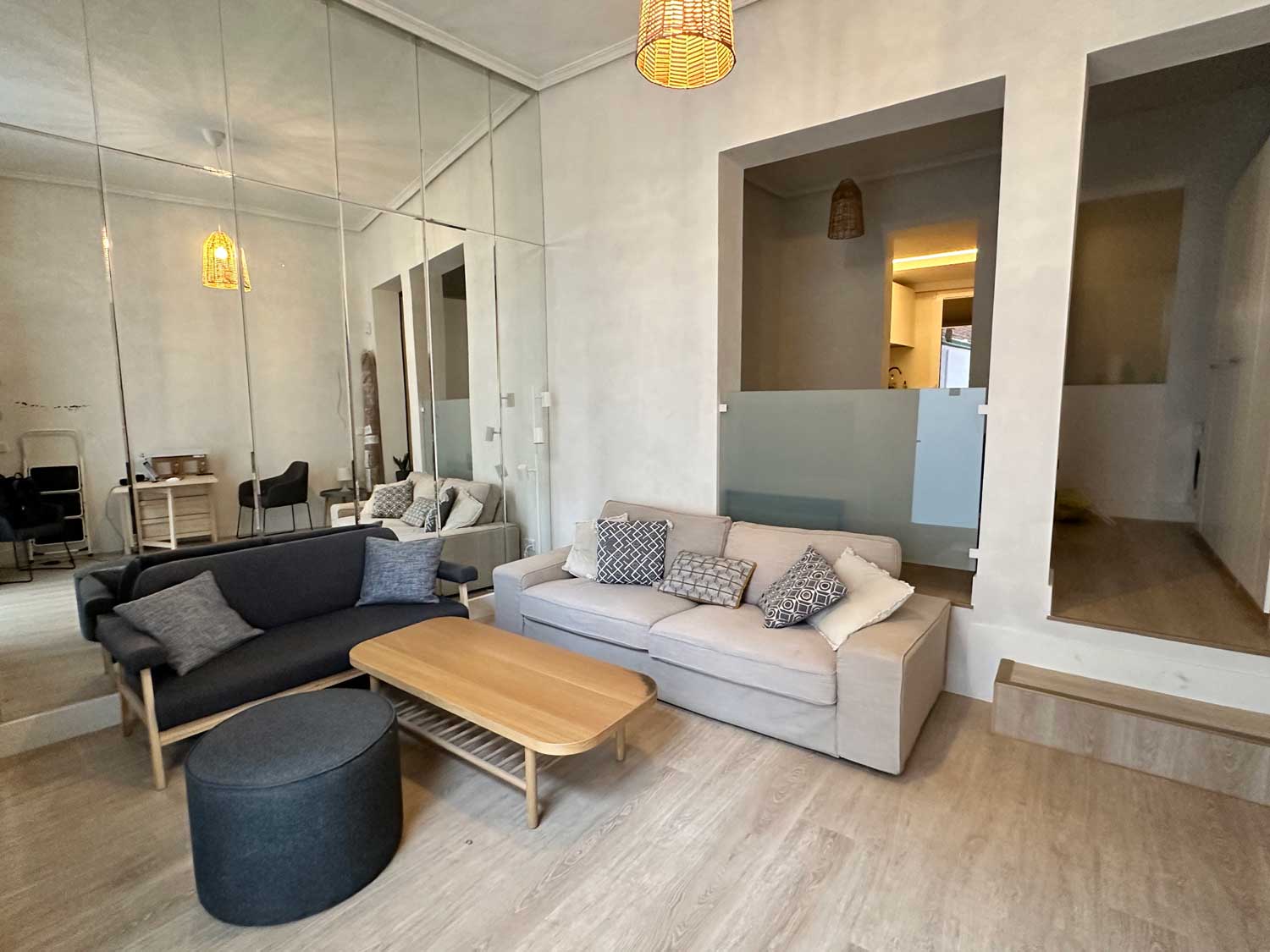 Una moderna sala de estar con un sofá gris con cojines estampados y un loveseat a juego. Una mesa de centro de madera ocupa el centro. Una pared de espejos refleja el espacio y hay luces cálidas en el techo.