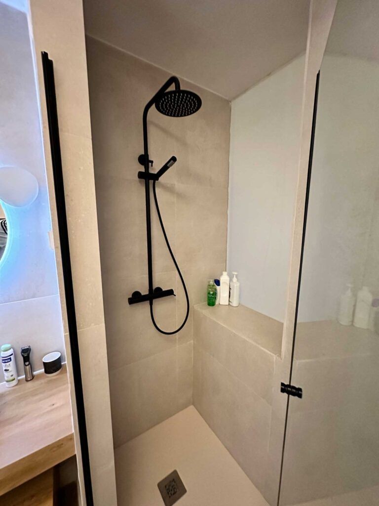 Una ducha moderna con grifería negra, incluyendo un cabezal de ducha tipo lluvia y un rociador de mano. La ducha tiene azulejos beige y puertas de cristal transparente. Los artículos de aseo están cuidadosamente ordenados en la esquina. Una encimera de madera es parcialmente visible.