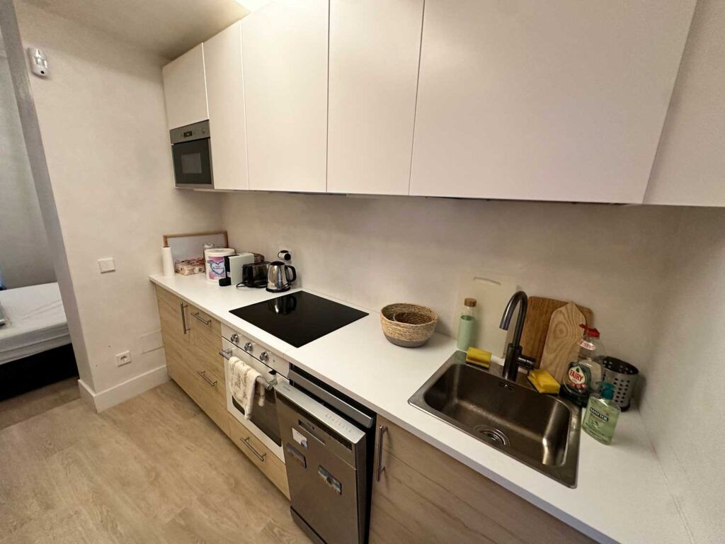 Cocina moderna con gabinetes blancos y piso de madera clara. Cuenta con fregadero, placa eléctrica, horno y microondas. La encimera alberga diversos artículos, como una tetera, tablas de cortar y utensilios de cocina. La puerta da a otra habitación.