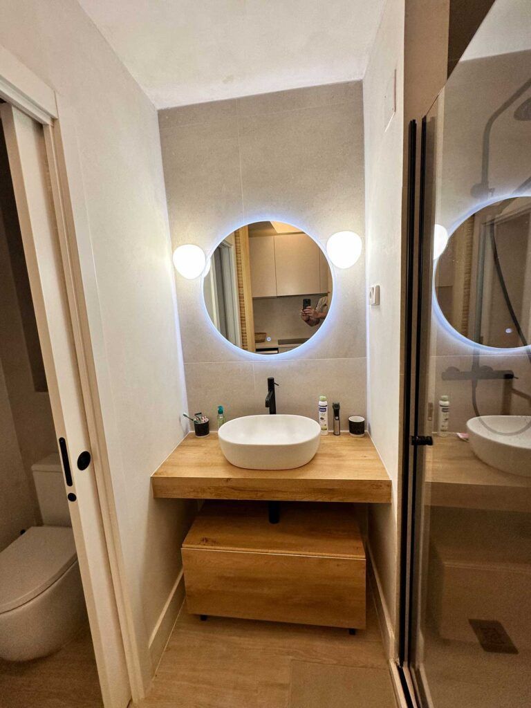 Un baño moderno con un espejo redondo iluminado sobre un tocador de madera, lavabo blanco sobre encimera y grifería negra. Dos apliques esféricos flanquean el espejo. A la derecha hay una mampara de ducha de cristal y una puerta corredera da acceso al aseo.