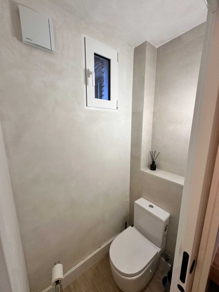 Un baño pequeño con un inodoro blanco y un portarrollos en la pared izquierda. Hay una ventana estrecha sobre el inodoro y una pequeña repisa a la derecha con un difusor de varillas. Las paredes están pintadas de un color claro.