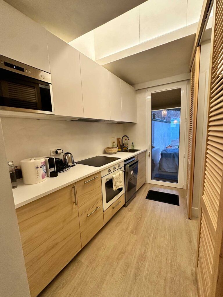 Cocina moderna con gabinetes de madera clara, encimeras blancas y electrodomésticos de acero inoxidable. Una puerta da a un patio. Hay pequeños electrodomésticos y utensilios de cocina en la encimera, y una puerta de persiana a la derecha.