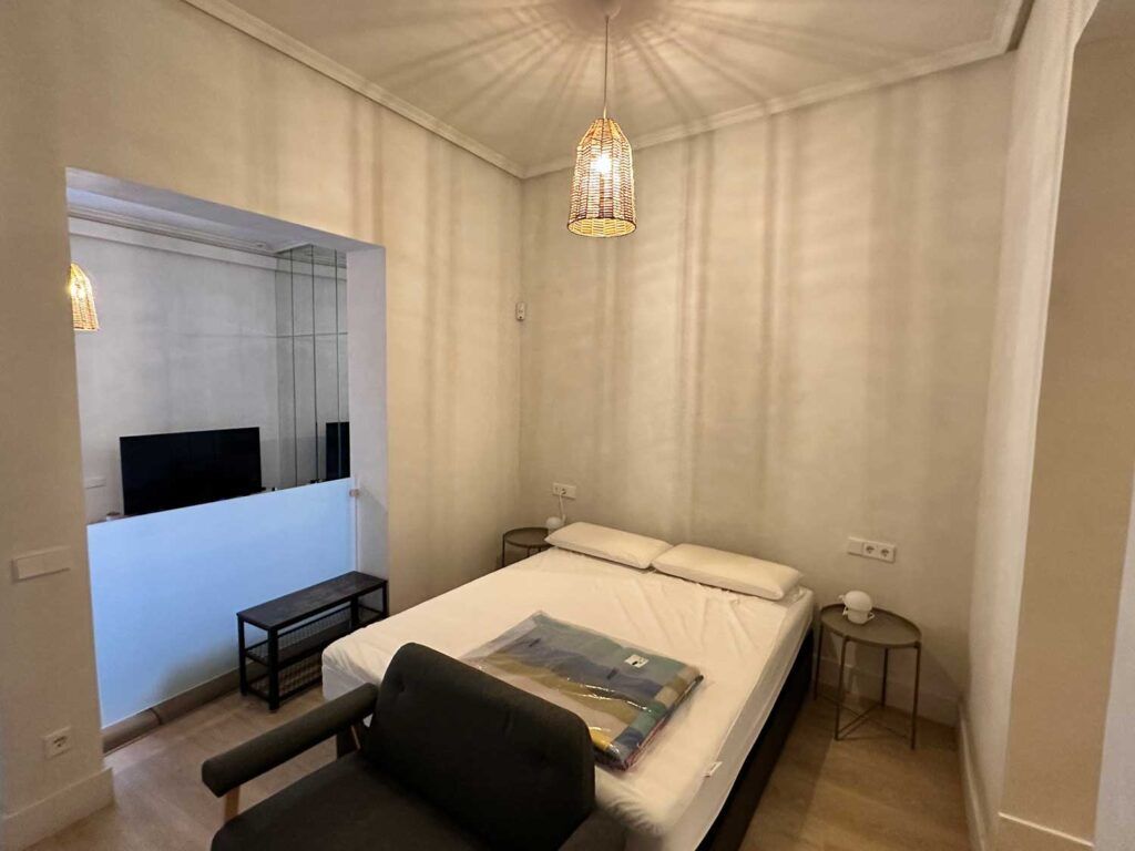 Un dormitorio minimalista con una cama, dos almohadas, un sofá negro a los pies y un televisor sobre un mueble. La habitación, de colores neutros, está iluminada por una lámpara colgante, lo que crea un ambiente acogedor.
