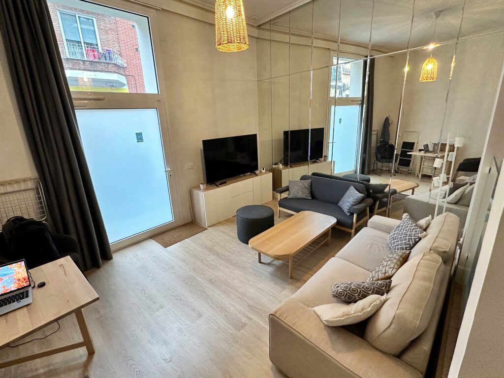 Una sala de estar moderna con un sofá beige, una mesa de centro de madera y un sillón gris frente al televisor. Una pared de espejos refleja el espacio. Una computadora portátil se encuentra sobre un escritorio de madera cerca de una ventana con cortinas oscuras. Lámparas colgantes cuelgan del techo.