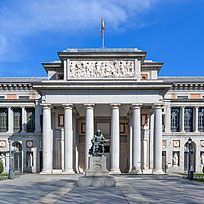 Vista frontal del Museo del Prado en Madrid, con arquitectura clásica, altas columnas, una estatua central y tallas intrincadas sobre la entrada. El edificio se recorta contra un cielo azul despejado.