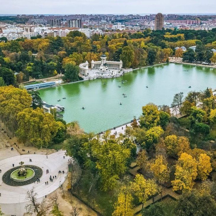 Vista aérea de un gran parque con un lago verde rodeado de árboles otoñales. Hay pequeñas embarcaciones en el lago, y una estructura neoclásica de piedra se alza en un extremo. El paisaje urbano se extiende al fondo bajo un cielo despejado.